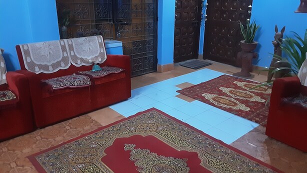 Un canapé rouge recouvert d’une housse en dentelle, un tapis à motifs et une porte bleue ornée de motifs métalliques décoratifs.