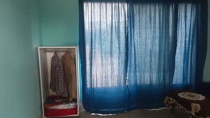 Une pièce aux murs de couleur turquoise, une armoire remplie de vêtements, un lit recouvert d’une courtepointe à motifs floraux et un rideau bleu.