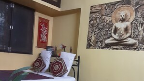 Une pièce comprenant un lit, un climatiseur fixé au mur, un tableau décoratif représentant un Bouddha, un coussin rouge et blanc, un tapis à motifs et une plante en pot.