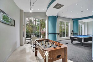 Un área recreativa moderna en el interior, con mesa de fútbol, mesa de billar y ventanas amplias que ofrecen vistas al exterior.