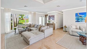 Un vaste salon comprenant un grand canapé, une table basse et un coin repas avec une table en verre. Un tableau orne le mur et de grandes fenêtres offrent une vue sur l’extérieur.