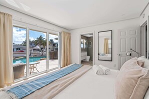 A hotel room with a bed, a large window with a view of a pool and palm trees, a bathroom with a mirror, and a door.