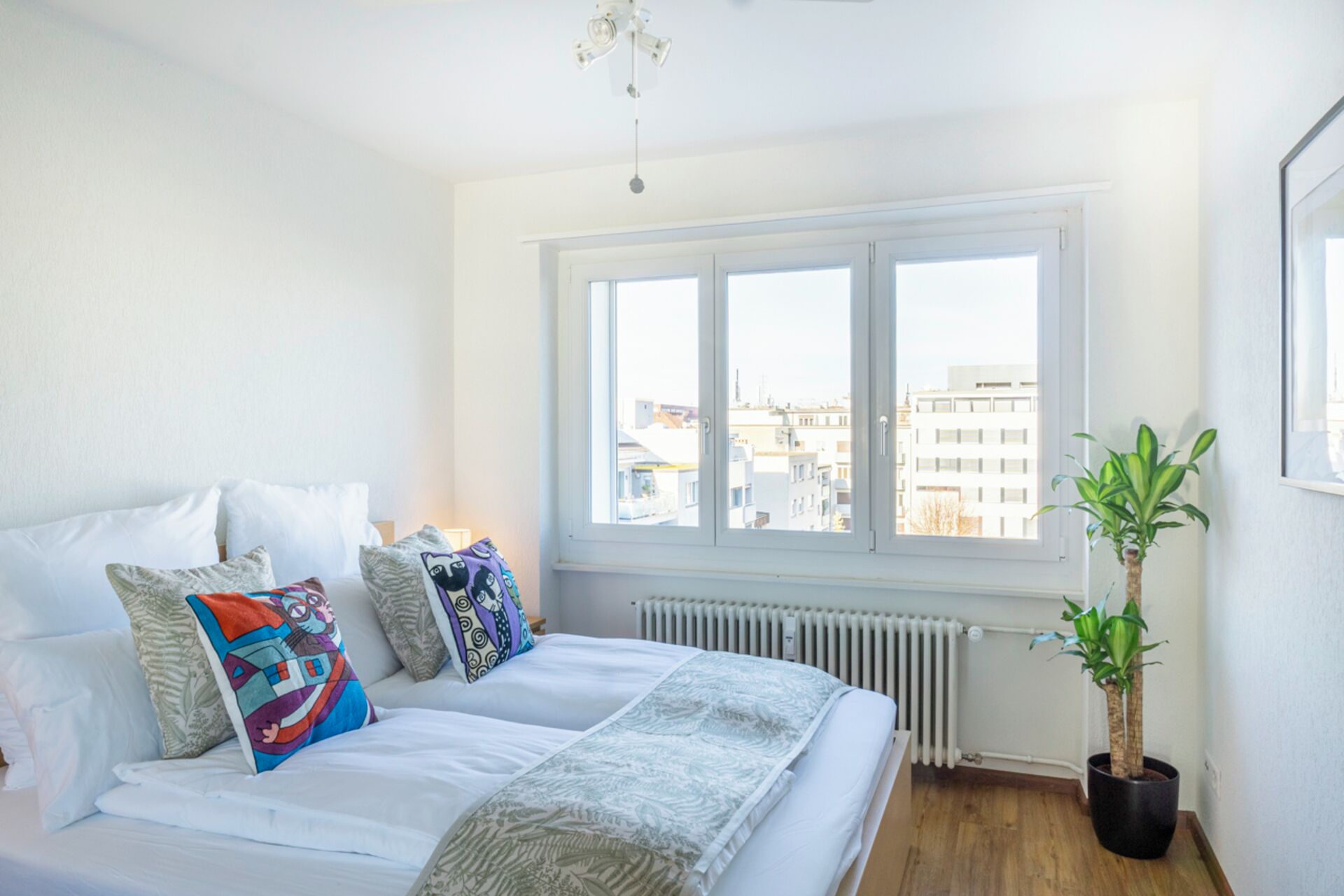 Ein Schlafzimmer mit großem Fenster, einem Bett mit weißer Bettwäsche, mehreren Kissen, einem Heizkörper und einer Topfpflanze.