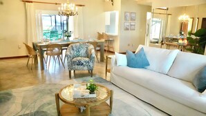 Una sala de estar con un sofá blanco, una mesa redonda y un sillón estampado. Se observa un comedor con una mesa y sillas al fondo.