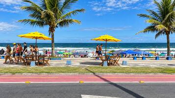 Um calçadão à beira-mar com palmeiras, guarda-sóis e pessoas caminhando.