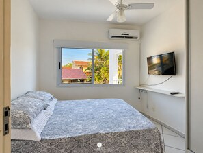 Un dormitorio con cama, televisor, aire acondicionado y ventana con vistas a edificios y árboles.