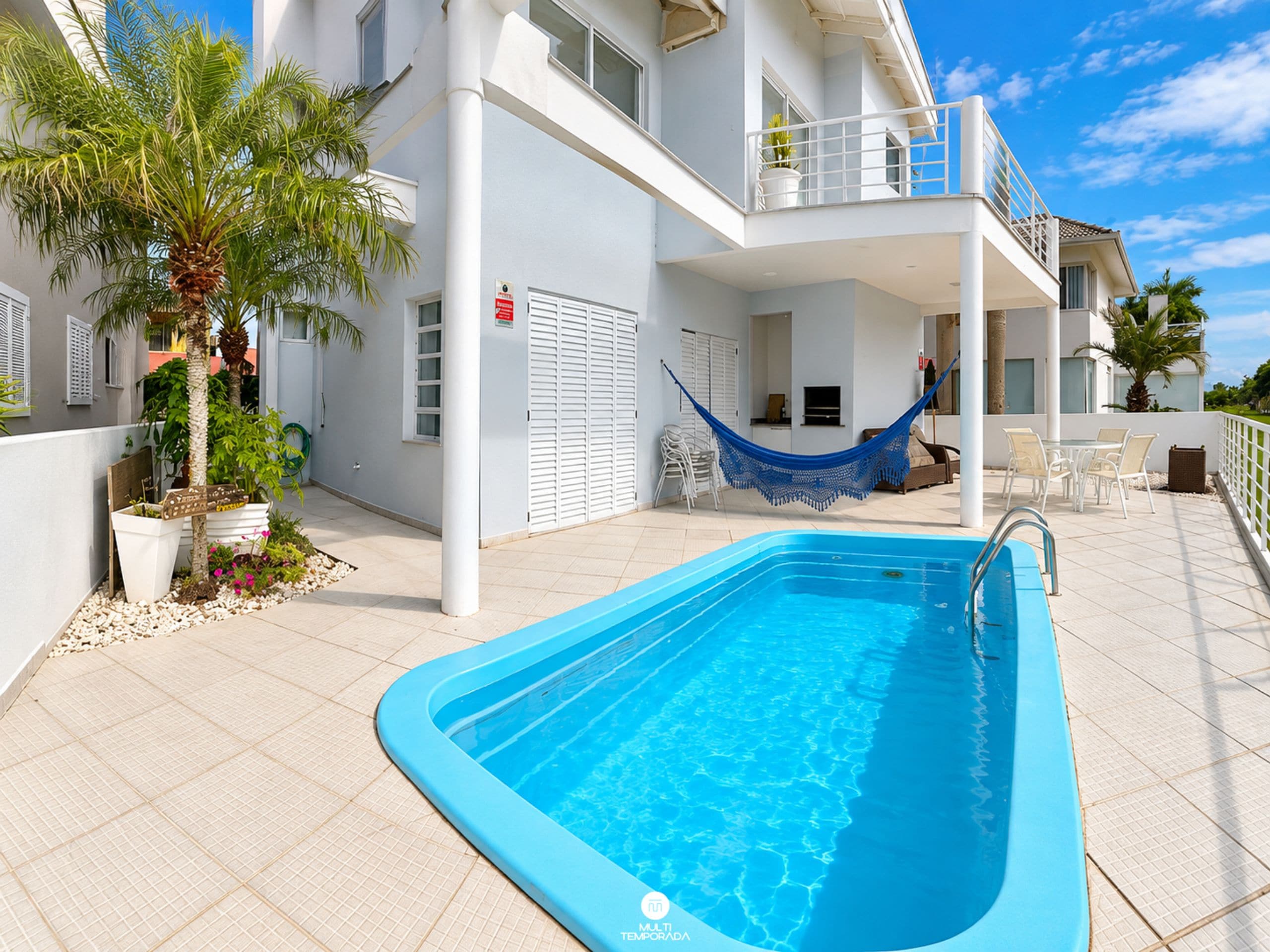 Una piscina con borde azul, rodeada por un edificio blanco con balcones y plantas en macetas.