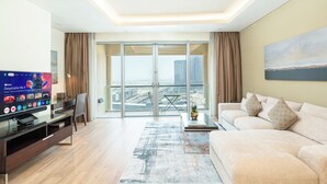 A modern living room with a flat-screen TV, a sofa, a coffee table, and a view of a cityscape through large glass doors. - PVH - Emaar Fashion Avenue - 1BR (Dubai)