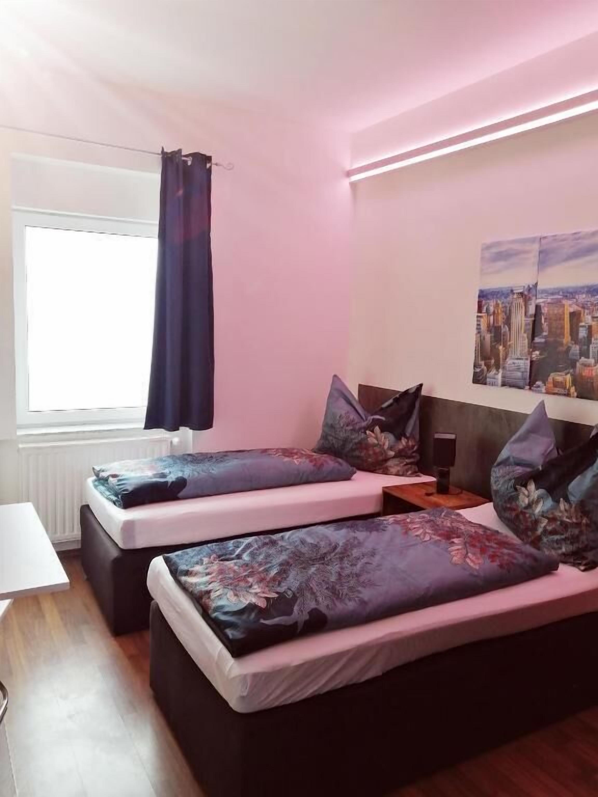 A bedroom with two beds, a window with a curtain, a cityscape painting on the wall, and a small table with a speaker.