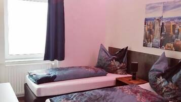 A bedroom with two beds, a window with a curtain, a cityscape painting on the wall, and a small table with a speaker.