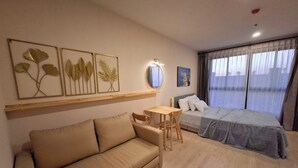 A hotel room with a sofa, a small table, a bed with a blue cover, a window with curtains, and wall art.