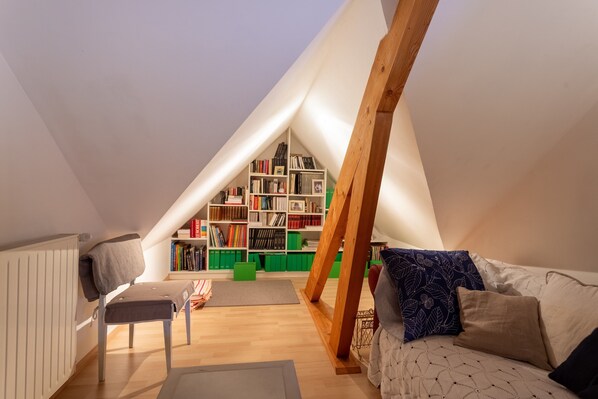 A cozy attic room with a sloped ceiling, a wooden chair, a sofa, a bookshelf, and a radiator.