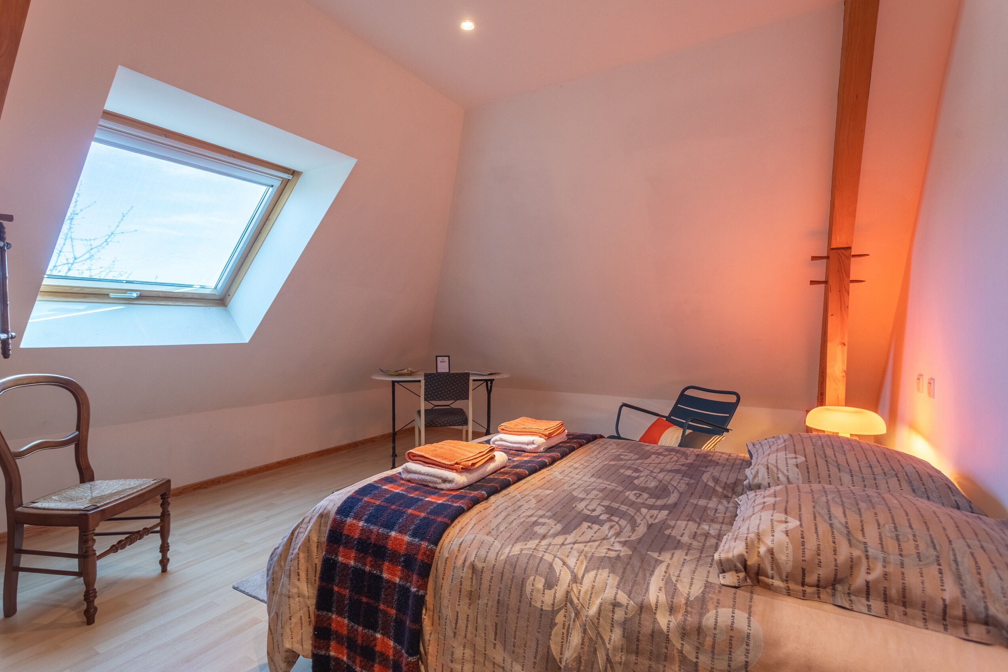 Une chambre avec un puits de lumière, un lit recouvert d’une couvre-lit à motifs, une chaise, un bureau et une lampe.