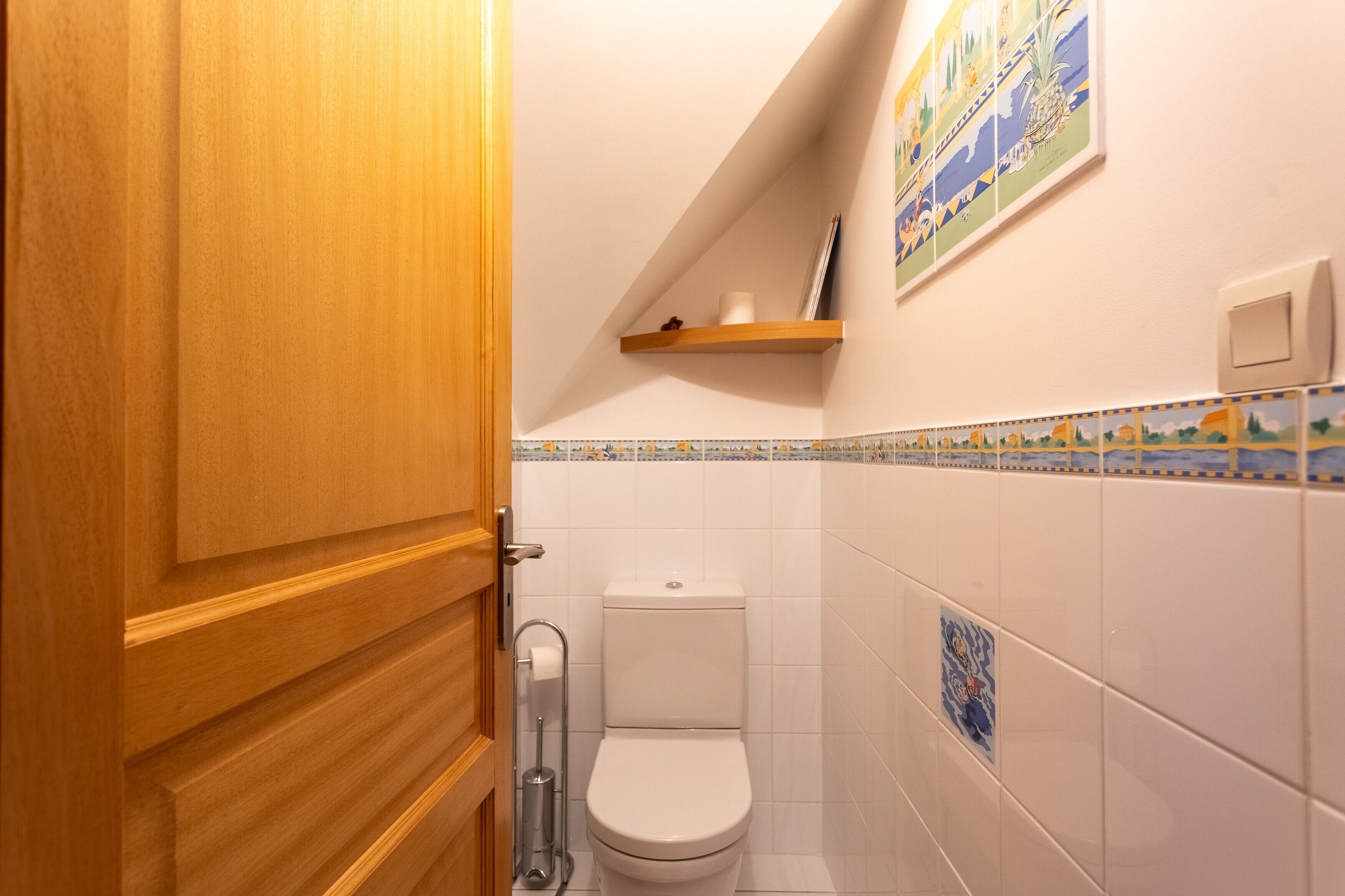 Un baño con puerta de madera, inodoro blanco, repisa con un libro y velas, y pared con diseño azul y amarillo.