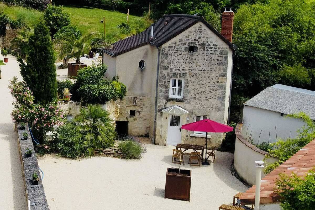 A stone building with a tiled roof, surrounded by greenery and a paved area with outdoor furniture.