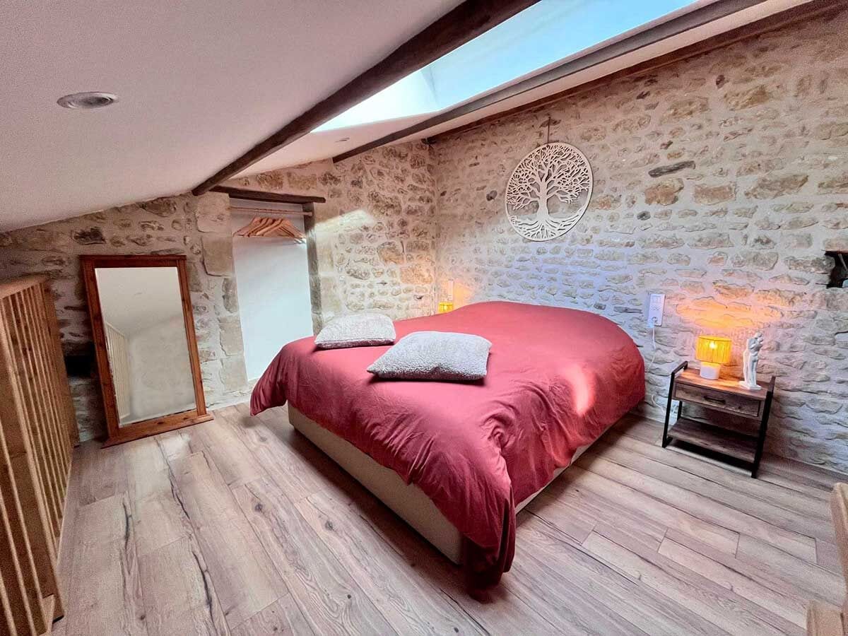 A bedroom with a stone wall, a bed with a red bedspread, a wooden floor, a nightstand with a lamp, and a mirror.