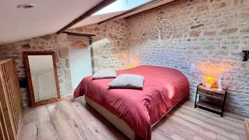 A bedroom with a stone wall, a bed with a red bedspread, a wooden floor, a nightstand with a lamp, and a mirror.