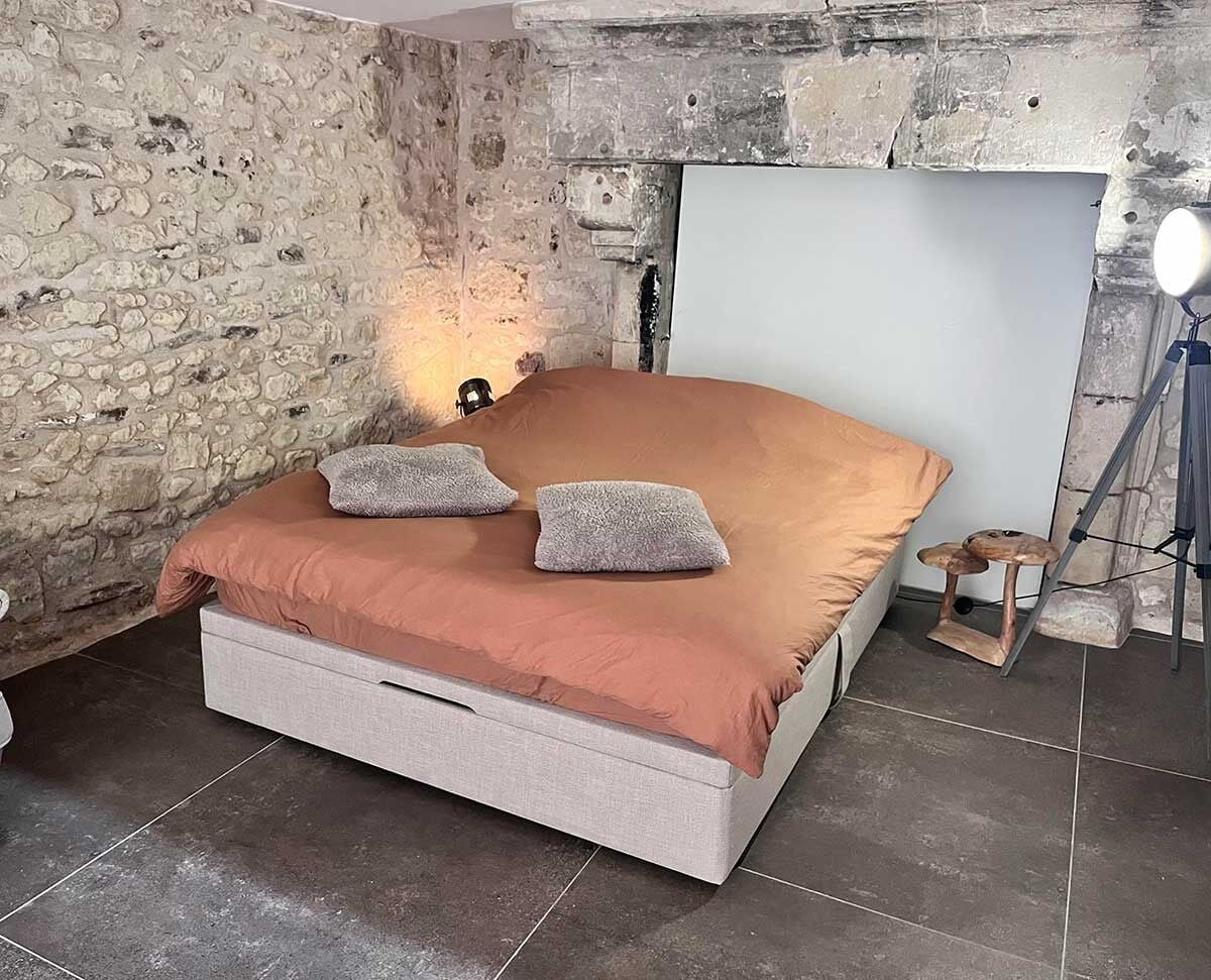 A bed with a headboard against a stone wall, a bedside table with a lamp, and a small stool.