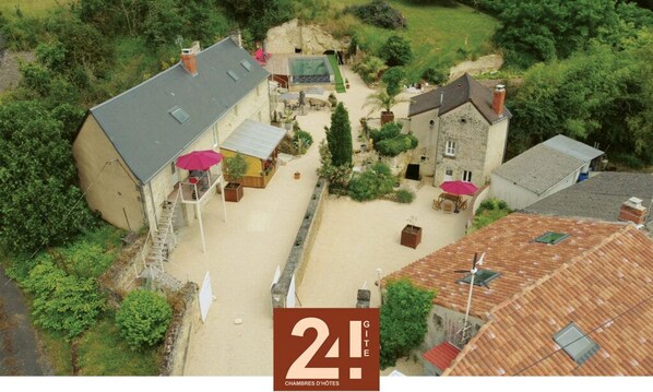 An aerial view of a property with multiple buildings, including a main house with a balcony and a smaller structure with a red umbrella. There are outdoor seating areas and a garden with various plants. - Le Gîte 24 (Beaumont Saint-Cyr)
