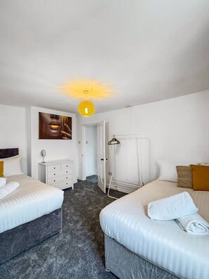 Habitación de hotel con dos camas, un tocador blanco, un espejo, un cuadro en la pared y una lámpara colgante.