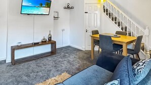 A living room with a sofa, a TV mounted on the wall, a dining table with chairs, and a staircase leading to another level.