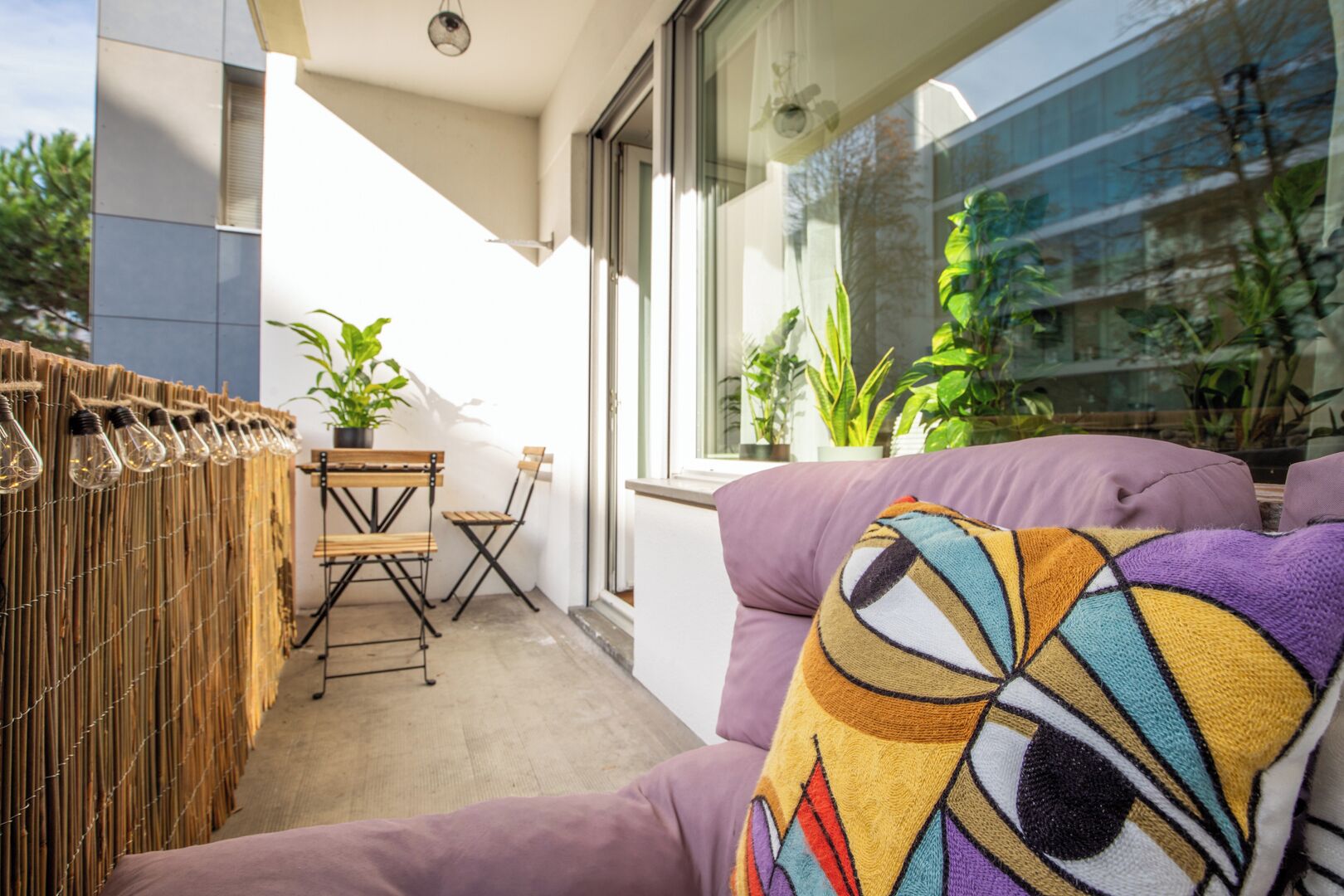 Ein Balkon mit gewebter Trennwand, einem Holztisch, zwei Stühlen und einem violetten Sofa mit farbigem Kissen. Übertöpfe mit Pflanzen und ein Fenster mit Blick auf Gebäude.