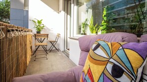 A balcony with a woven partition, a wooden table, two chairs, and a purple sofa with a colorful cushion. There are potted plants and a window with a view of buildings. - Special *PRIMERO PICASSO* Apartment (Basel)