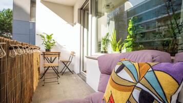Un balcone con un pannello divisorio intrecciato, un tavolo di legno, due sedie e un divano viola con un cuscino colorato. Sono presenti piante in vaso e una finestra con vista sugli edifici.