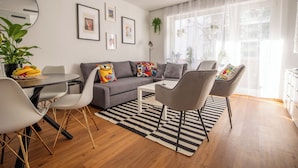A modern living room with a grey sofa, two armchairs, a coffee table, and a dining area with a white table and chairs. There are framed artworks on the wall and a large window with sheer curtains. - Special *PRIMERO PICASSO* Apartment (Basel)