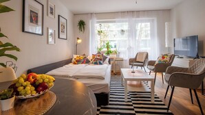 A modern hotel room with a bed, a dining table with fruit, a TV, and a large window with curtains. - Special *PRIMERO PICASSO* Apartment (Basel)
