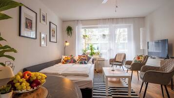 Ein modernes Hotelzimmer mit einem Bett, einem Esstisch mit Obst, einem Fernseher und einem großen Fenster mit Vorhängen.