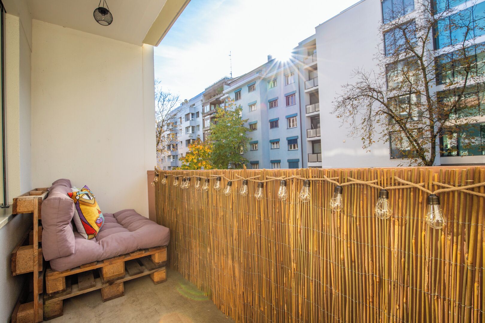 Un balcon doté d’un espace salon avec des coussins, d’une clôture en bambou et de guirlandes lumineuses.