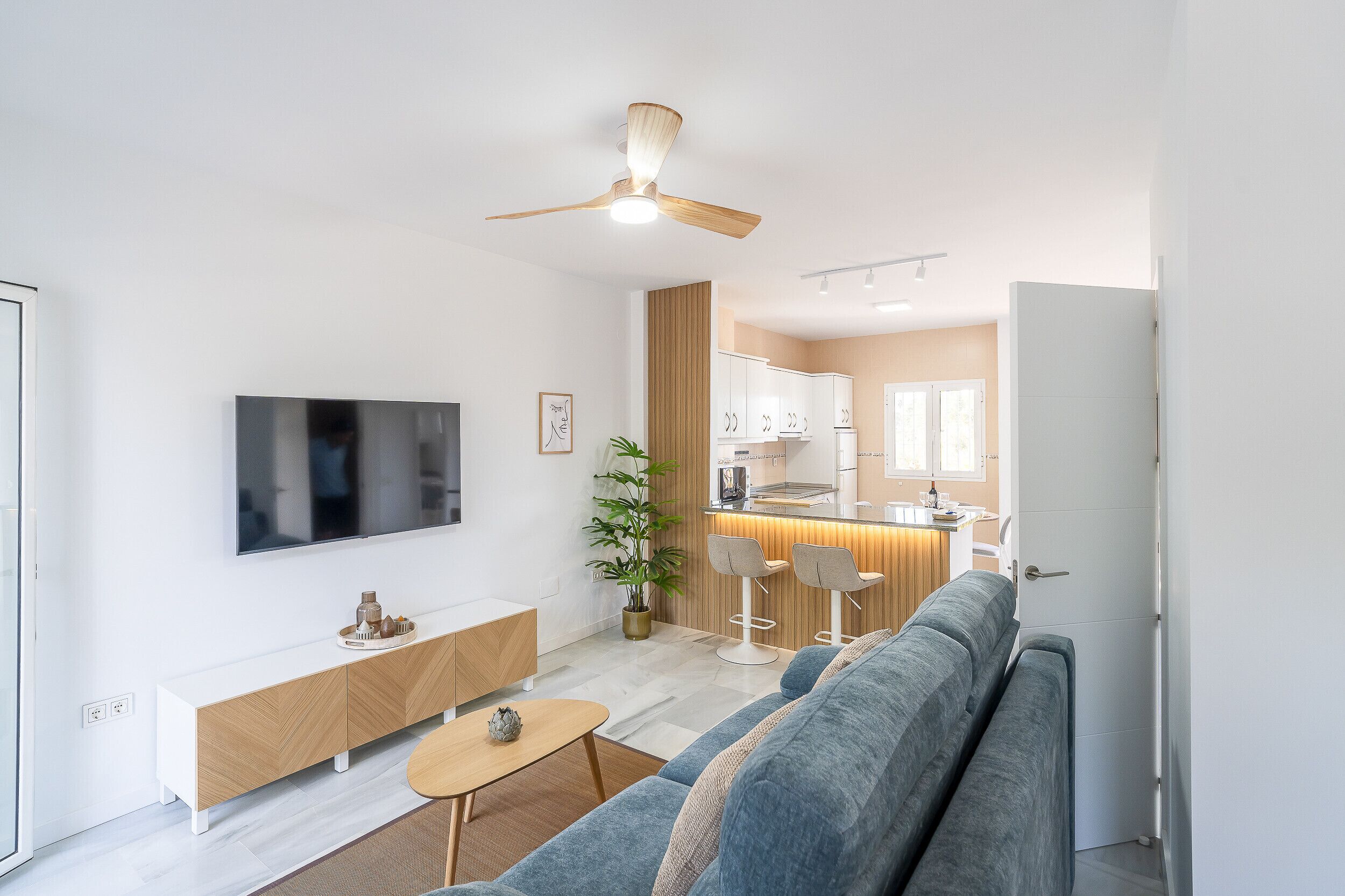 Ett modernt vardagsrum med en soffa, en tv och ett litet bord. En köksdel med en köksbänk och stolar syns genom en öppen dörr.