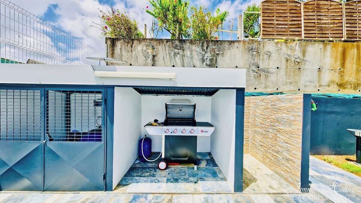 Eine kleine Außendusche mit Grill, Gasflasche und Schlauch, umgeben von einem Metallgitter und einer Betonmauer.
