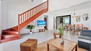 Un espacio habitable amplio con una escalera de madera, un televisor de pantalla plana, un comedor con mesa y sillas, y un sofá gris.