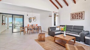 Un salón amplio con sofá gris, mesa de centro de madera y un comedor con sillas de madera y puerta de vidrio que da al exterior.
