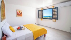 Un dormitorio con cama, una ventana con persianas, un cuadro y vista al mar.