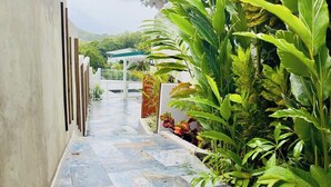 A narrow pathway with tiled flooring, bordered by lush greenery and a white fence.