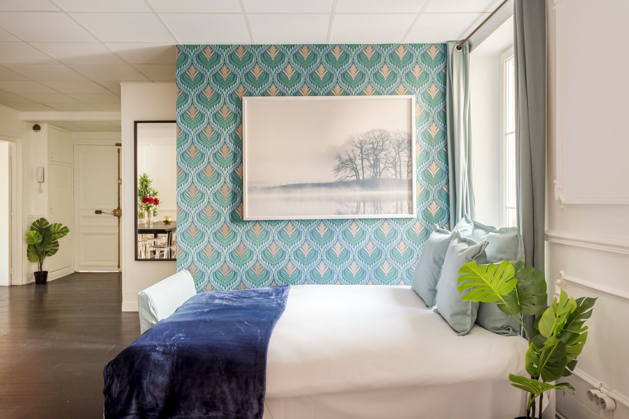 Een slaapkamer met behang met een patroon, een groot raam met gordijnen, een bed met kussens, en een plant op de vloer.