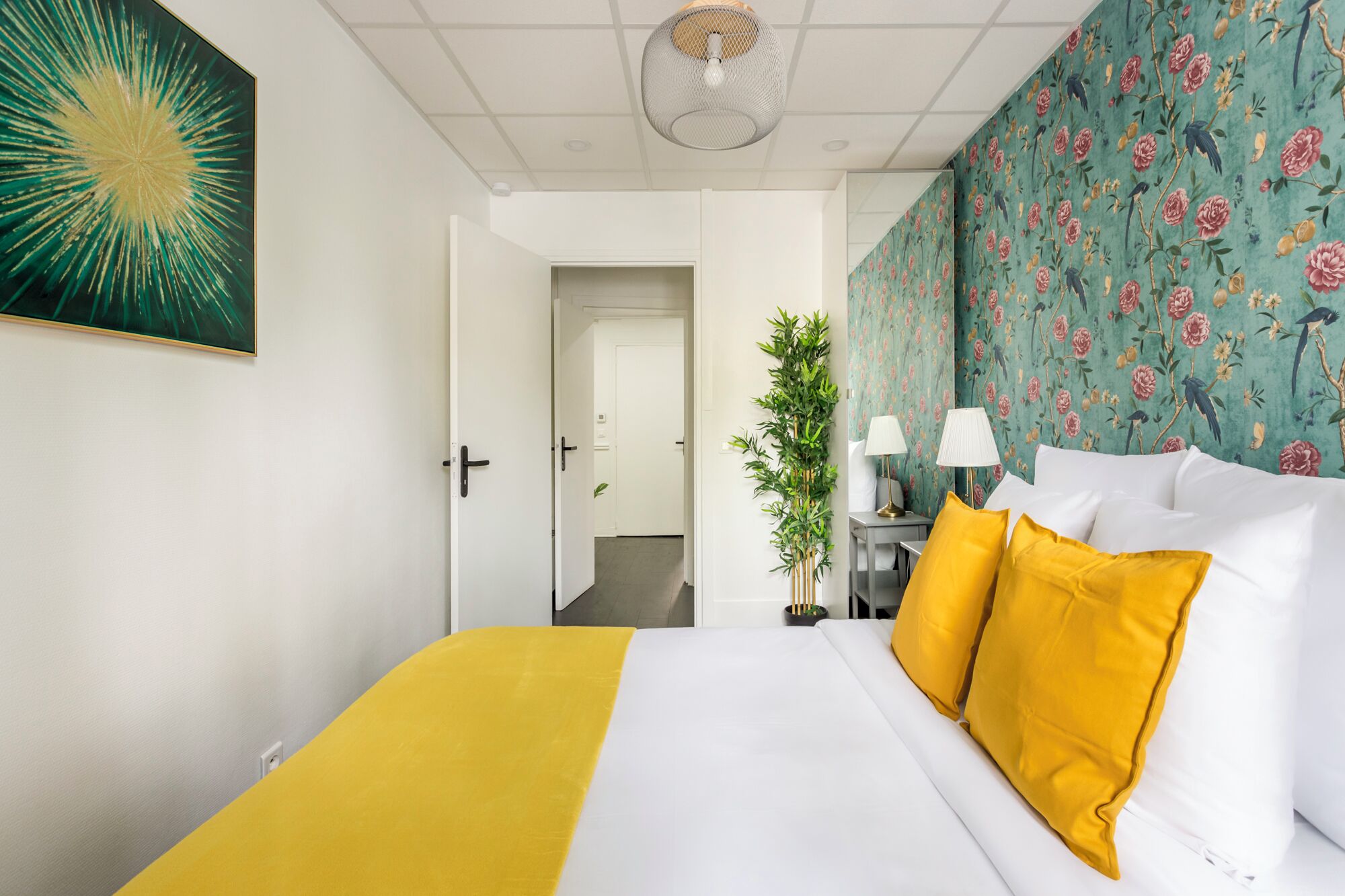 A hotel room with a bed, a large floral wallpaper, a green plant, and a painting on the wall.