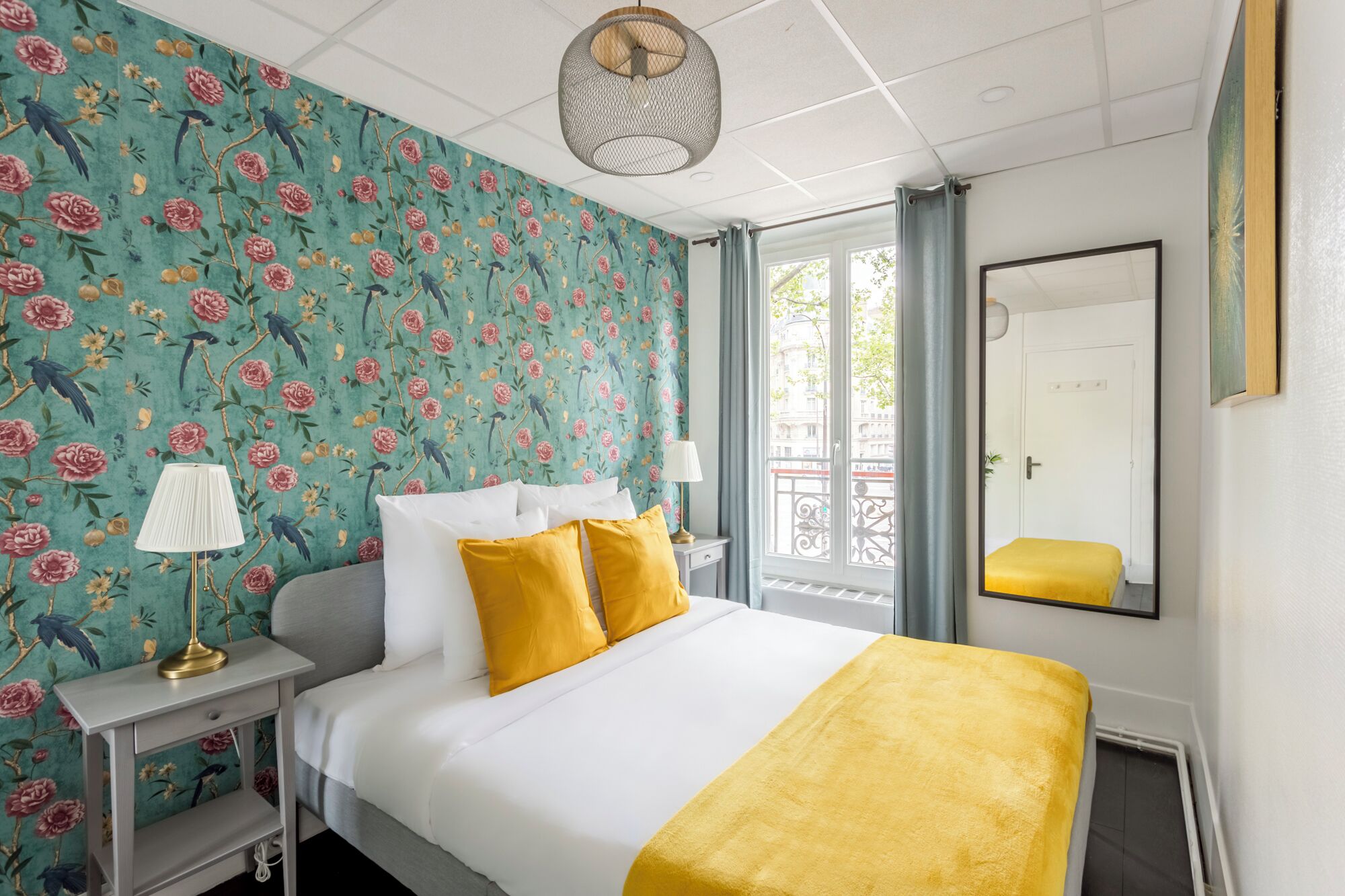 Habitación de hotel con cama, mesita de noche, lámpara, espejo y ventana con cortinas.