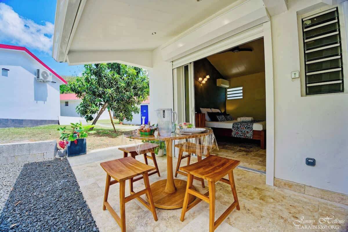An outdoor patio with a round wooden table and four chairs, a small garden area with potted plants, and a view of a building with a red roof.