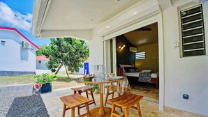 Um pátio externo com uma mesa de madeira redonda e quatro cadeiras, uma pequena área de jardim com plantas em vasos e vista para um edifício com telhado vermelho.