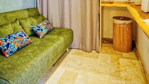 Um quarto pequeno e acolhedor com um sofá verde, duas almofadas coloridas, uma mesa de apoio de madeira com um vaso branco e um pequeno relógio, e um chão de azulejos bege.
