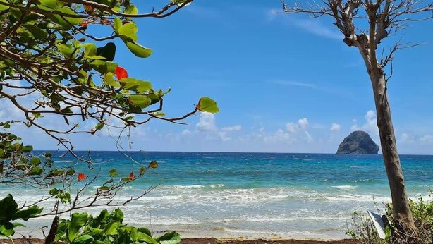 Uma praia com águas turquesa cristalinas, uma ilha rochosa ao longe e vegetação exuberante.