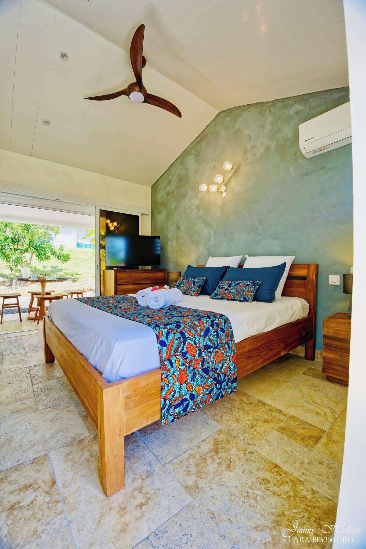 A bedroom with a wooden bed, a ceiling fan, a TV, and a view of the outdoors.