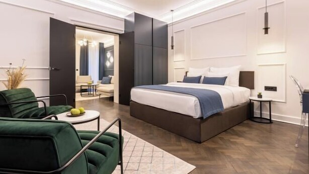 A modern bedroom with a large bed, a green armchair, a small round table, and a walk-in closet. - One Luxury Suites (Belgrade)