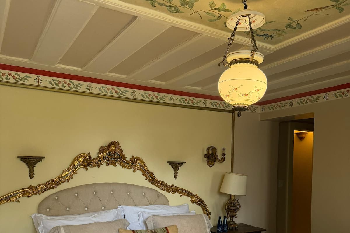 Une chambre d’hôtel avec un grand lit, une table de chevet en bois, une lampe et un plafond orné de motifs floraux.