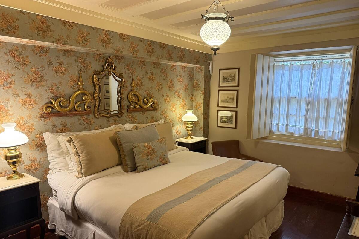 Une chambre à coucher avec un grand lit, une table de chevet avec une lampe, une chaise en bois, une fenêtre avec des rideaux et un mur recouvert d’un papier peint à motifs floraux et orné de tableaux encadrés.