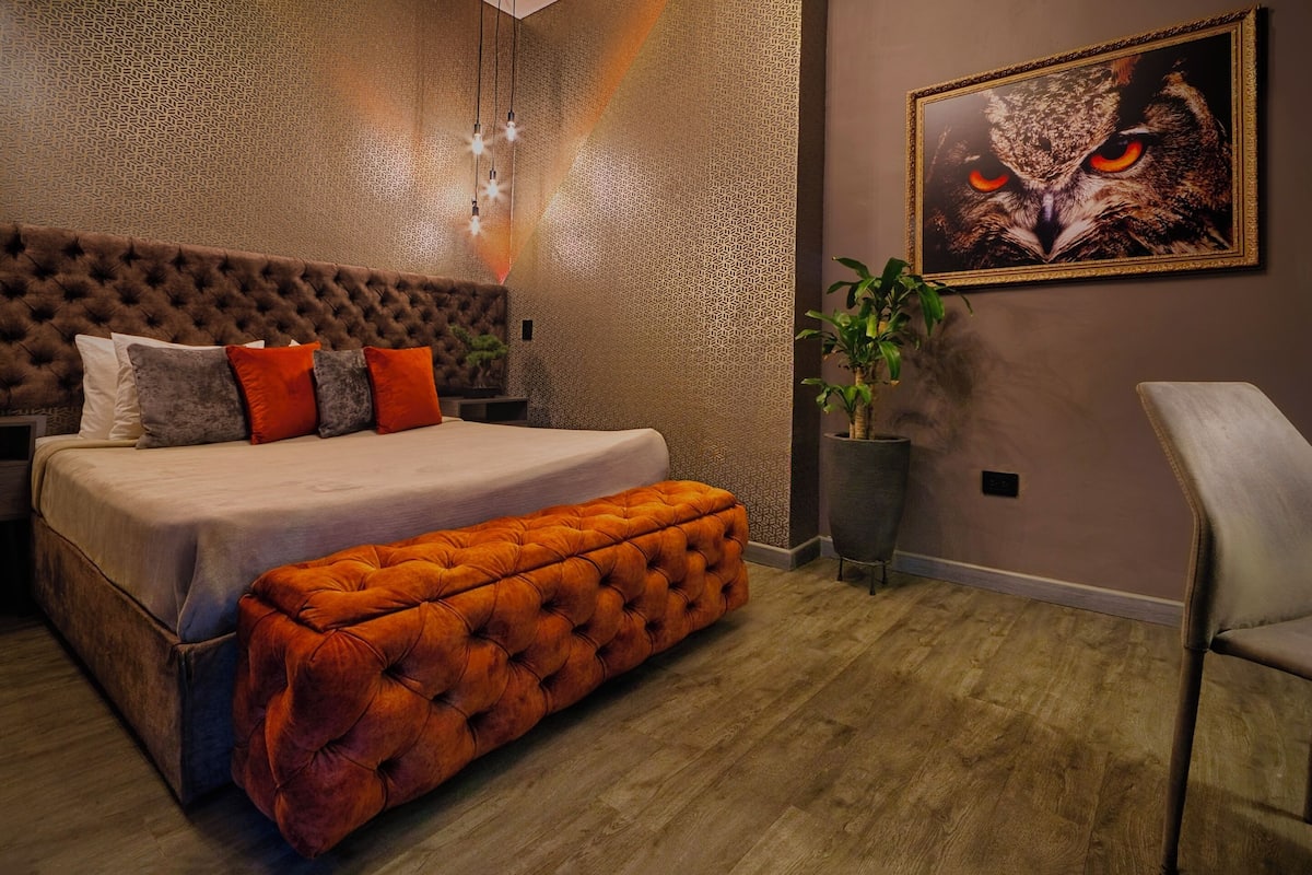 A bedroom with a tufted headboard, a large bed with multiple pillows, a bench, a framed owl artwork, a potted plant, and a chair.